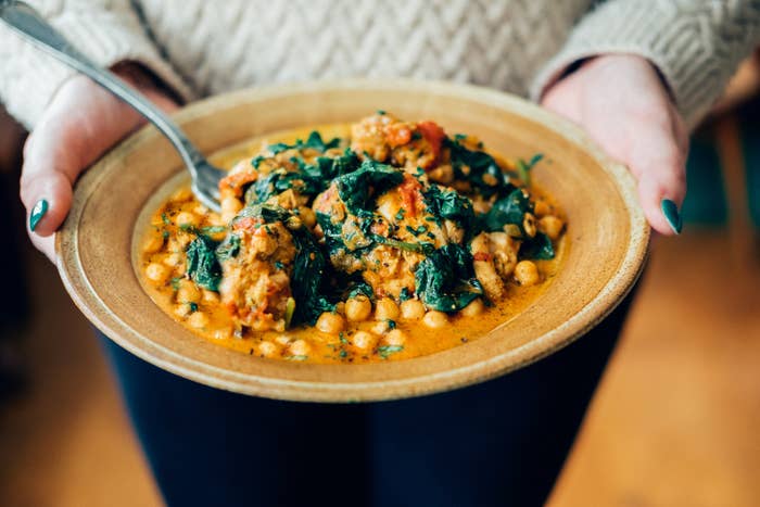 Hands holding a bowl of chickpea and spinach curry with tomatoes and spices.