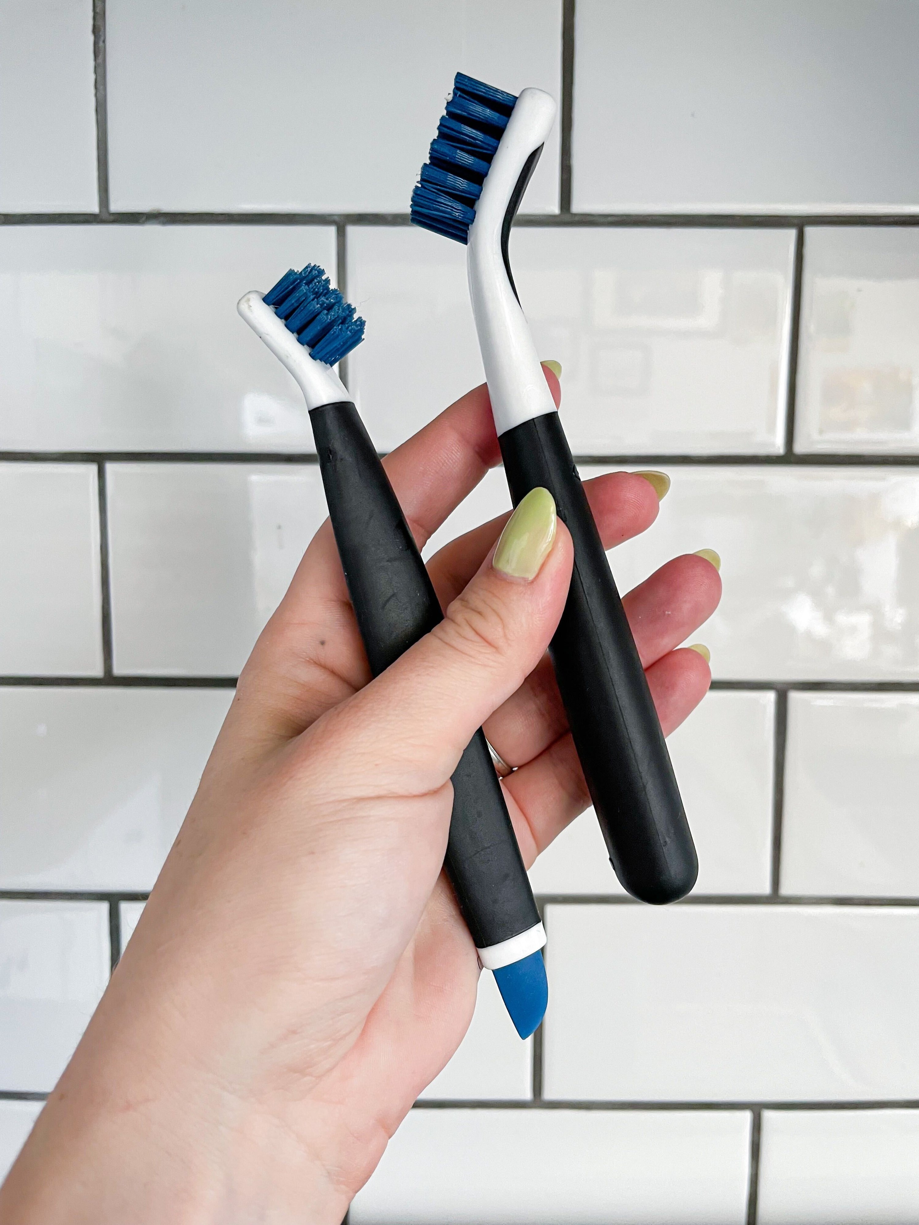 A person holding up the two mini scrubbing brushes against a subway tile backdrop