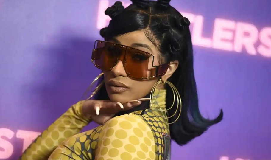 Cardi B in shades and a yellow outfit. She poses with an over the shoulder look.