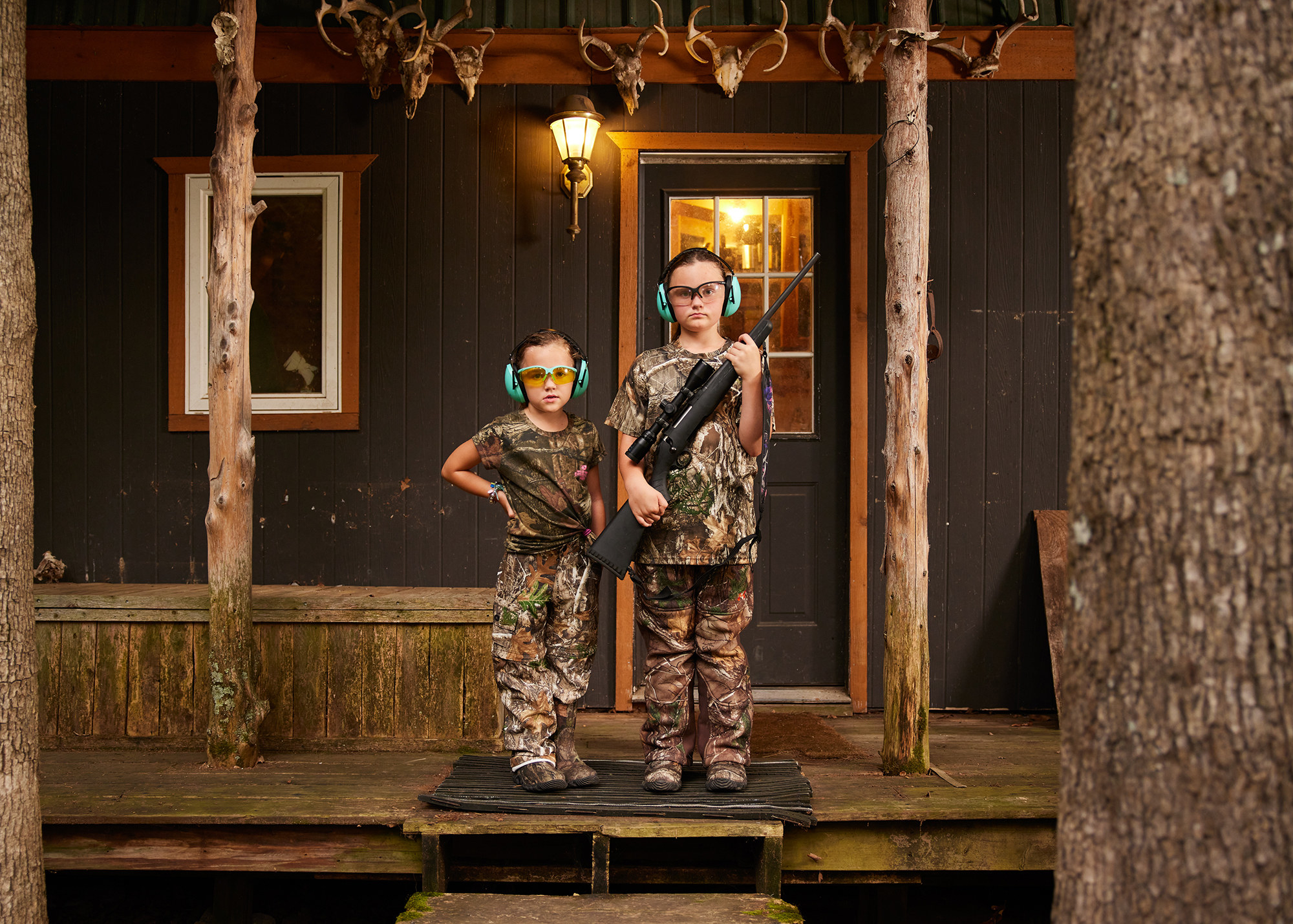 Two little girls in full camo with goggles and earmuffs carrying guns on the porch