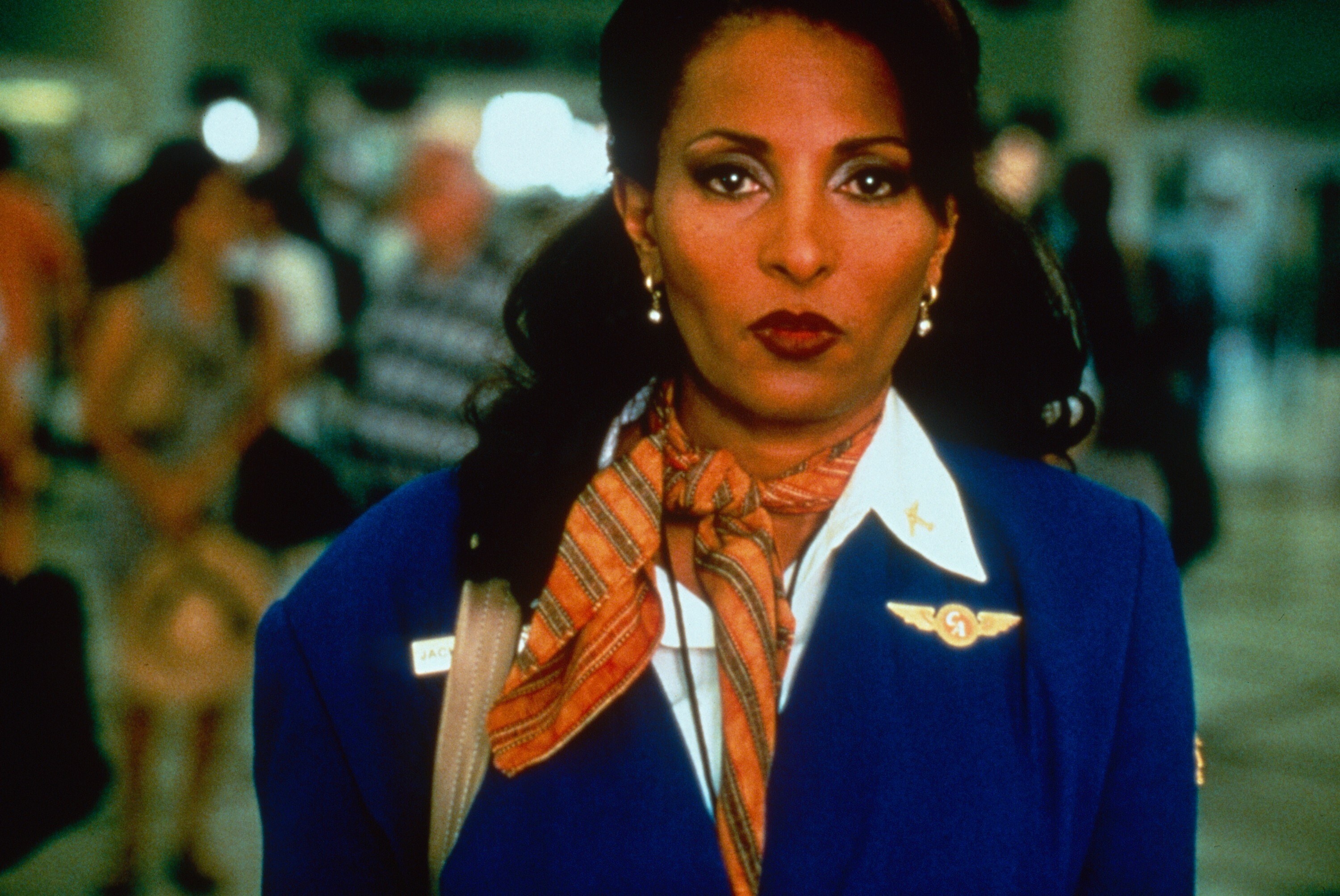 Pam Grier stands in an airport wearing a flight attendant uniform
