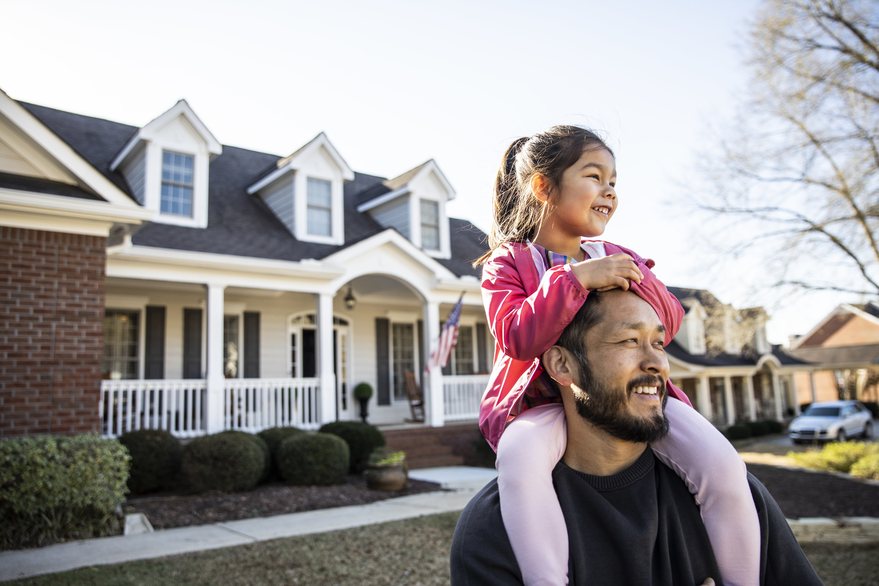 Dad giving young daughter a piggyback ride in front of a large suburban house