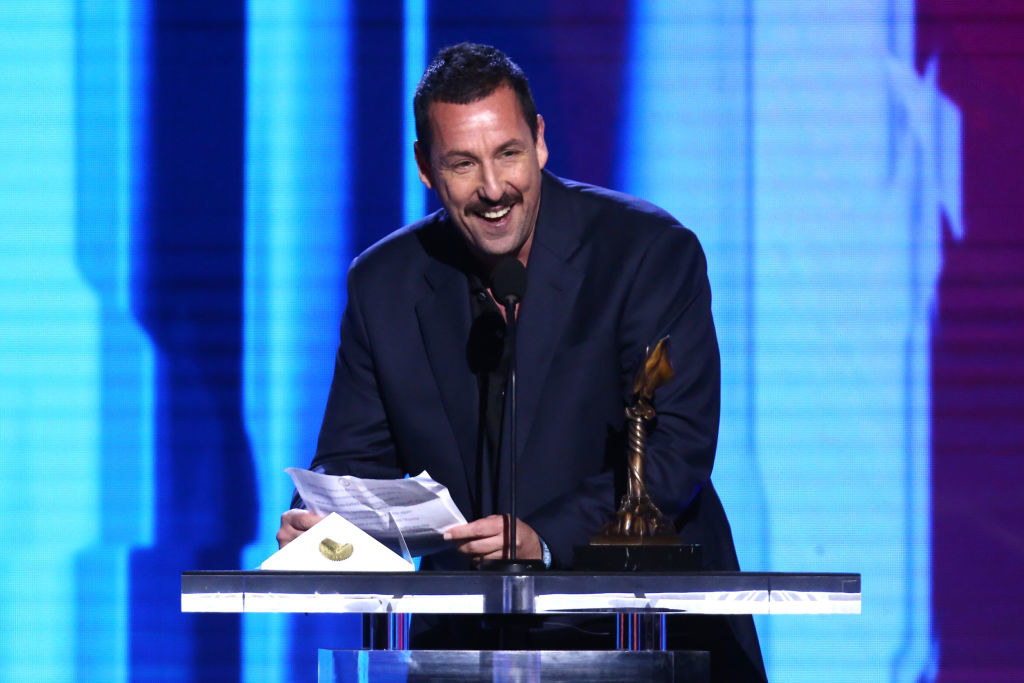 Adam Sandler speaking into a microphone and smiling