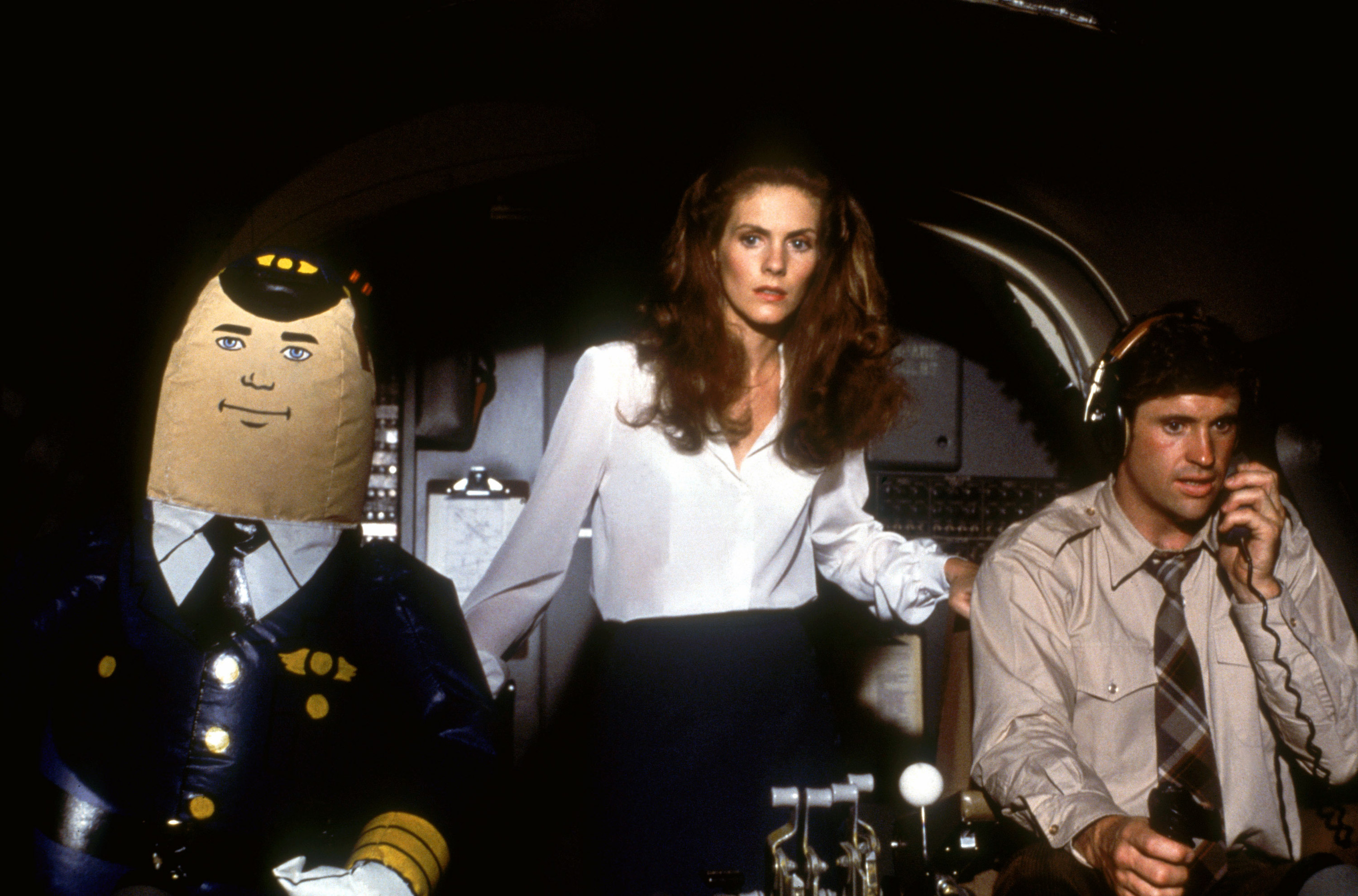 A flight attendant stands between a blow-up doll pilot and a real pilot in the plane cockpit