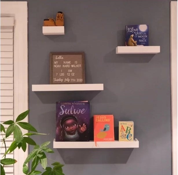 4 white floating shelves mounted on a white gray wall all holding books.