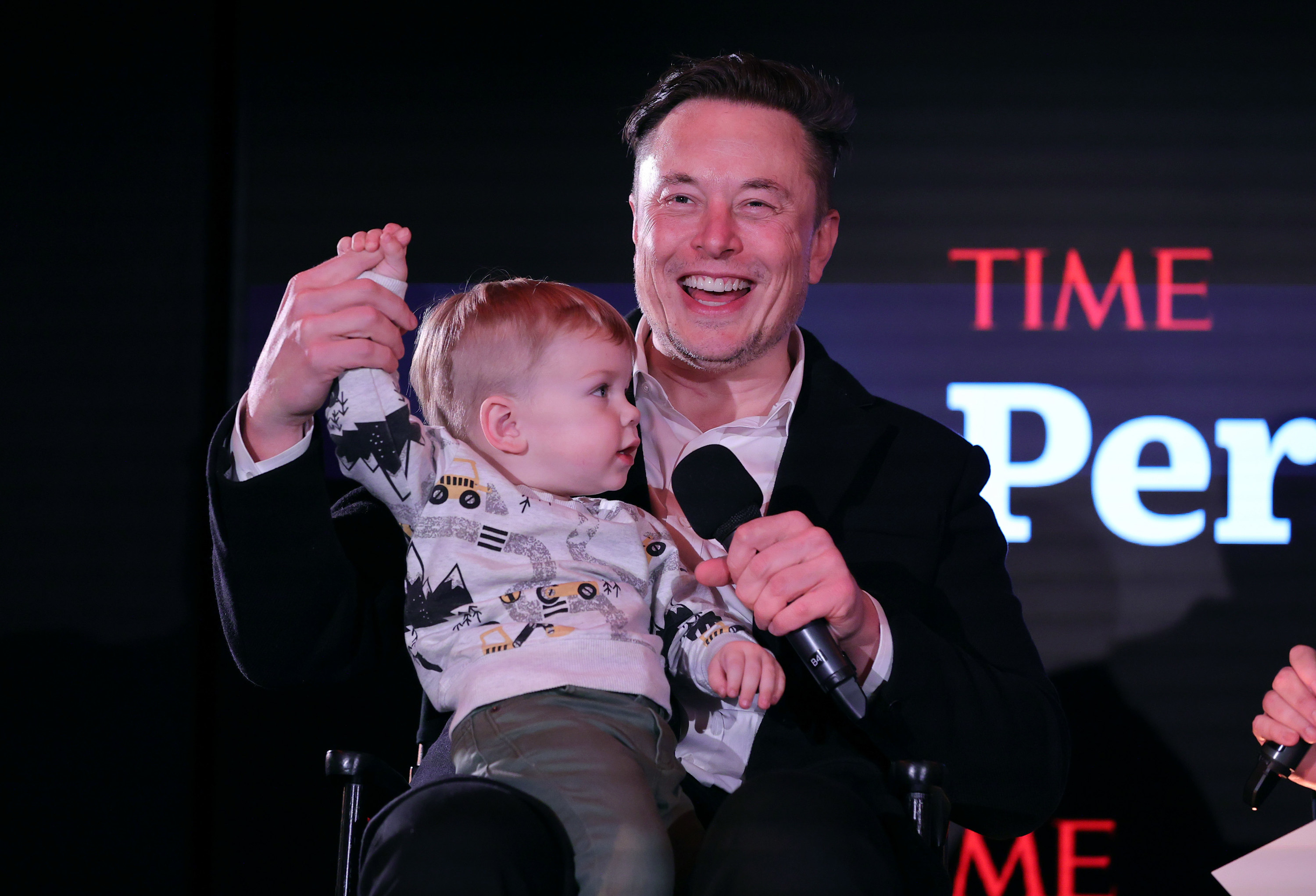 Elon smiles and holds his son on his lap at an event