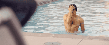 A man in a speedo coming out of a swimming pool as a lady looks at him.