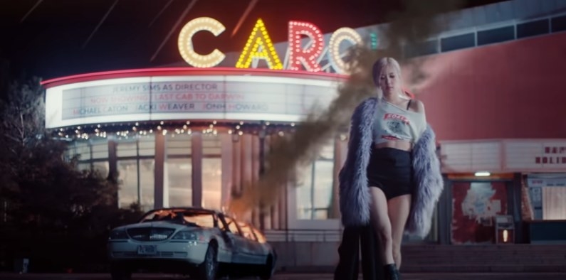 Rose walks away from a building at night, with is lit up with a neon saying spelling the word &quot;Carol&quot;