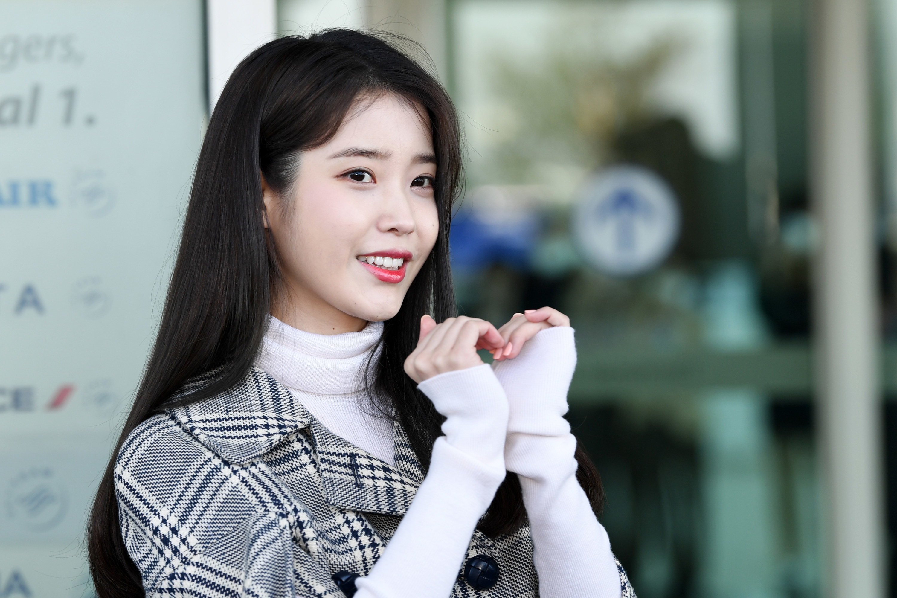 IU wears a turtleneck and plaid jacket; her hands form a heart shape in front of her, and she smiles with her long dark hair behind her shoulders