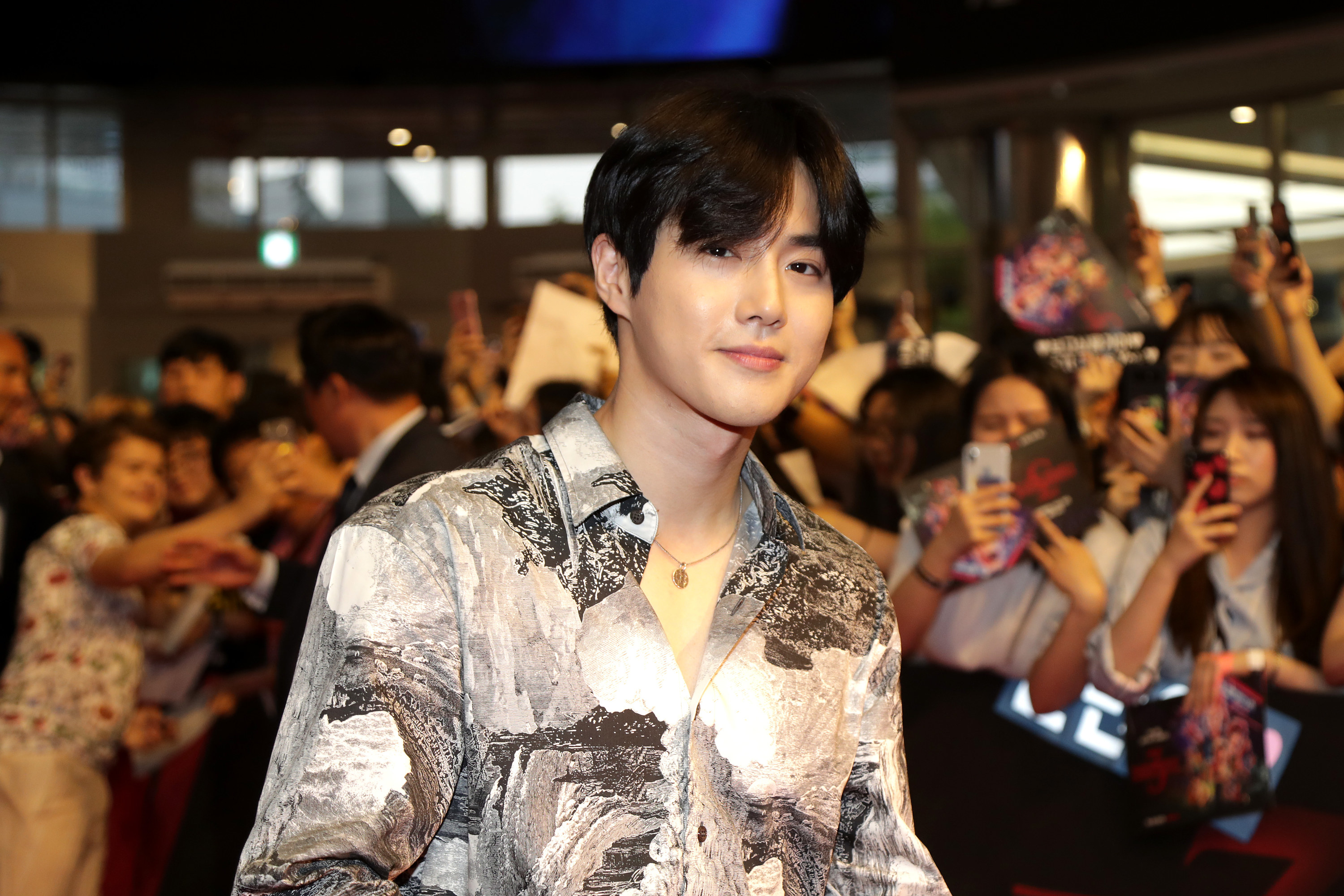 Suho smiles at the camera with floppy hair falling into his eyes; in the background, there are fans taking photos of him
