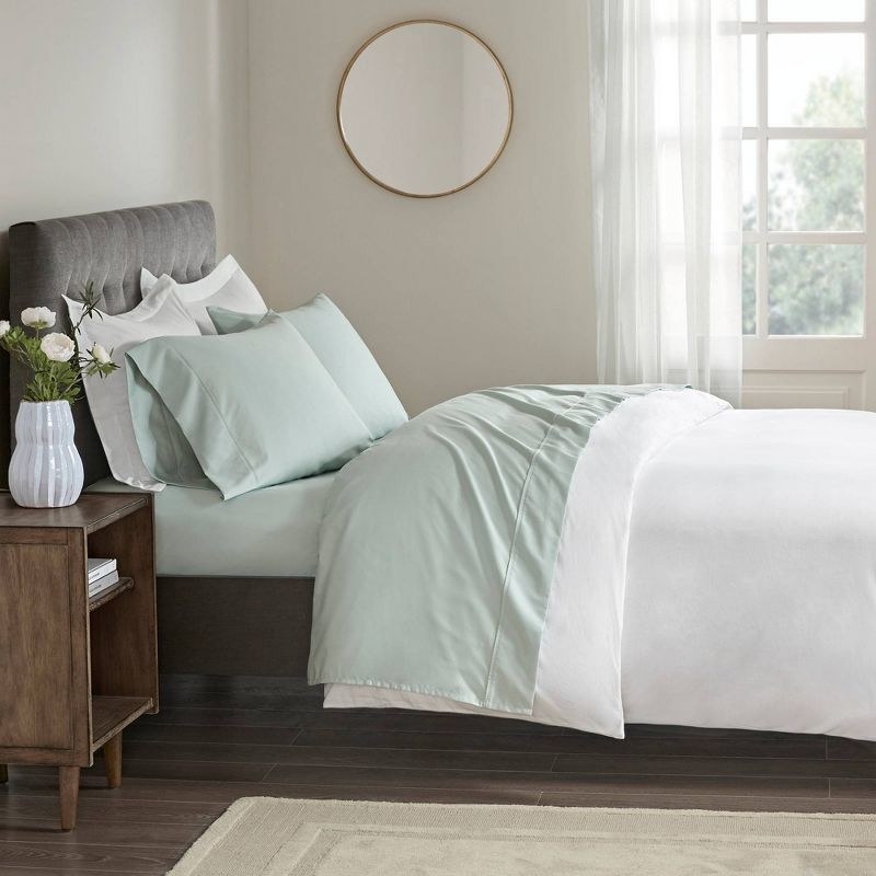 a made bed with green sheets in a bedroom with a nightstand, rug, and round mirror on the wall