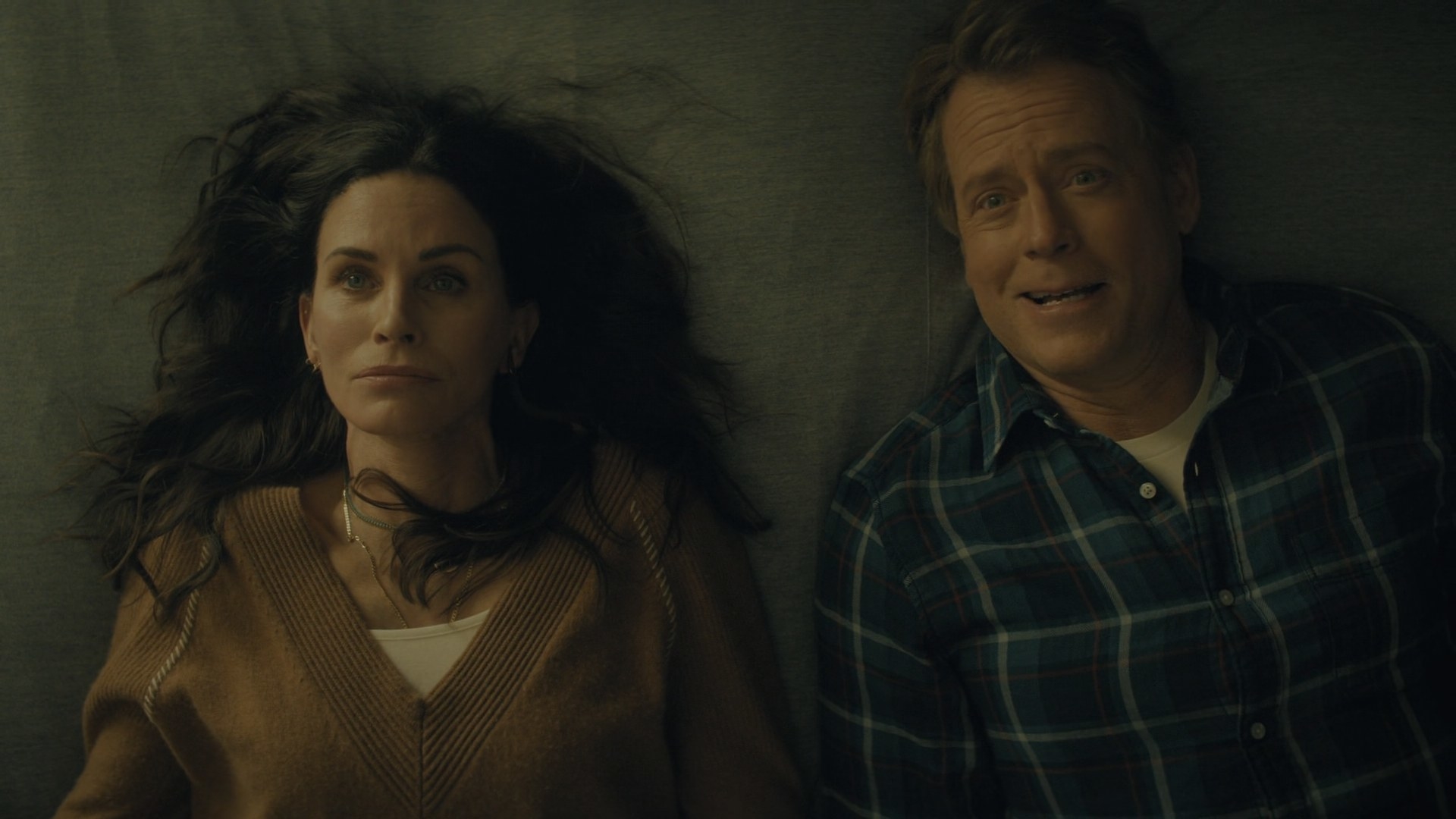 Courteney Cox and Greg Kinnear lie side by side on a bed