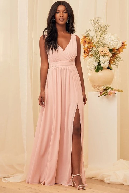 An image of a model wearing a blush surplice maxi dress that is sleeveless, has a V-back, and side slit