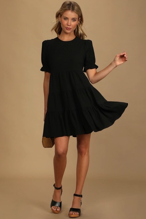 An image of a model wearing a black mini dress with puff sleeves, ruffle cuffs, and a relaxed bodice