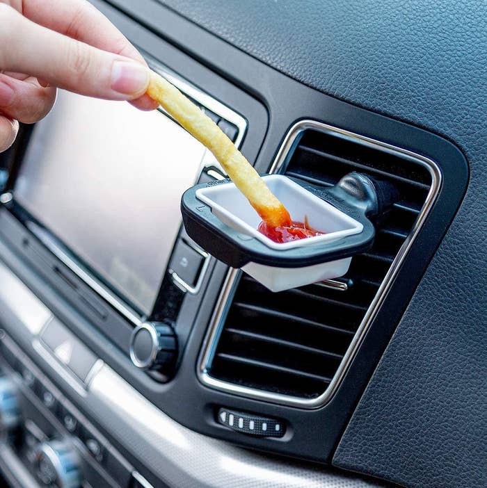 A person dipping a french fry into ketchup being held up by the holder in a car