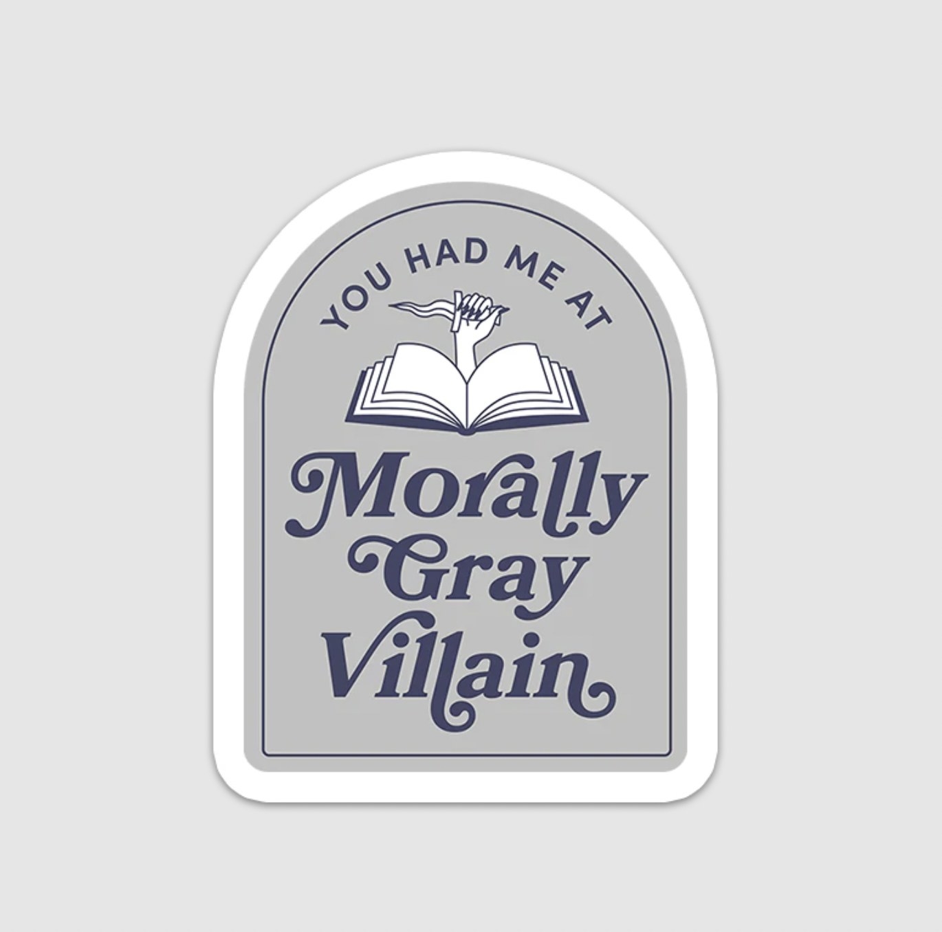 The tombstone shaped grey sticker with fancy words in dark grey reading &quot;you had me at morally gray villain&quot; with a hand holding a dagger and arising from open pages of a book