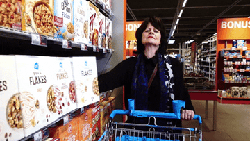 Woman pushing a cart down the aisle and knocking a row of cereal boxes into the cart