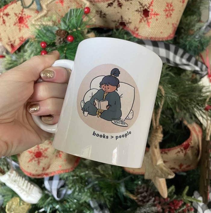 On the backdrop of a Christmas tree, someone holds the white mug with design of person sitting on pillows reading and message books&amp;gt;people underneath