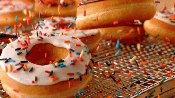 Rainbow sprinkles cascade down onto frosted donuts on a rack