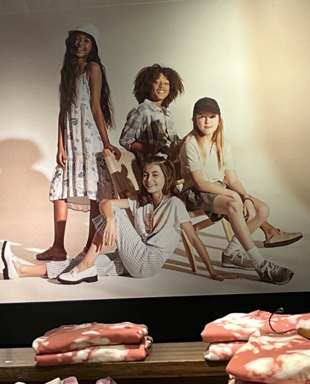 A group of smiling kids sit or stand together in a photo behind some folded clothes on a display