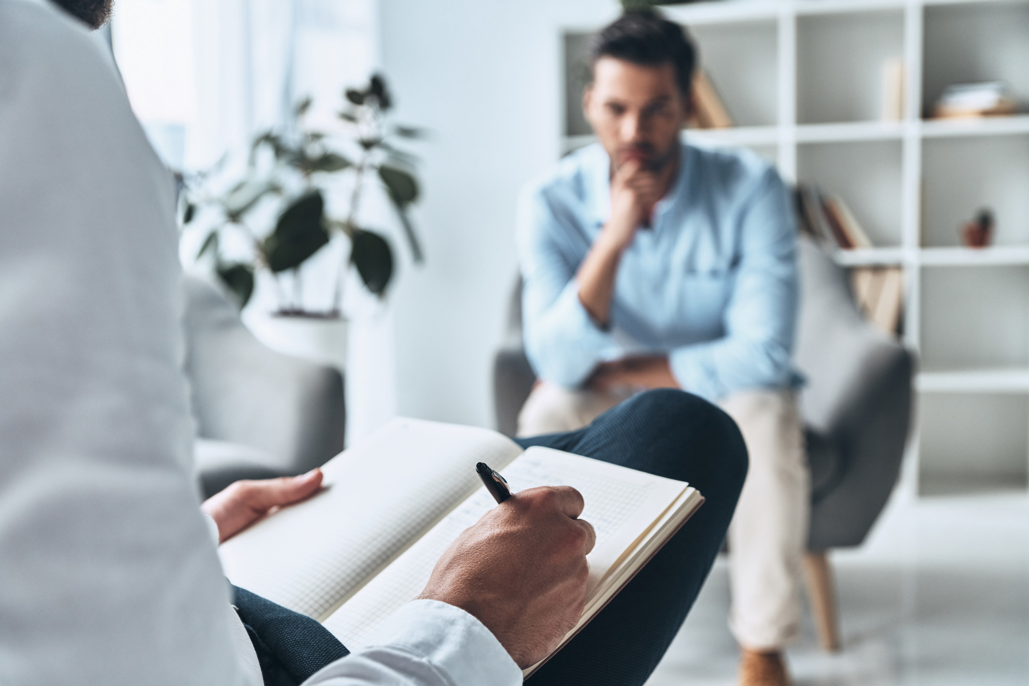 A man sits in a chair across from a therapist writing in a notebook