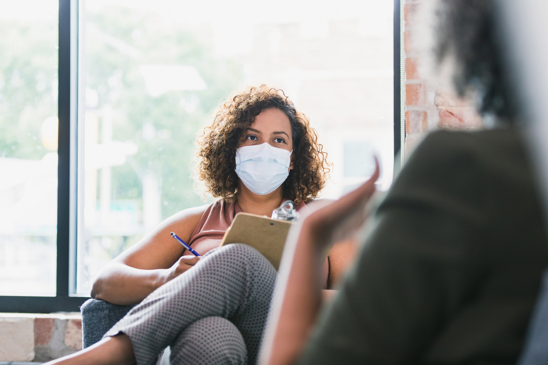 A therapist wearing a mask holds a clipboard and sits in a chair across from a client