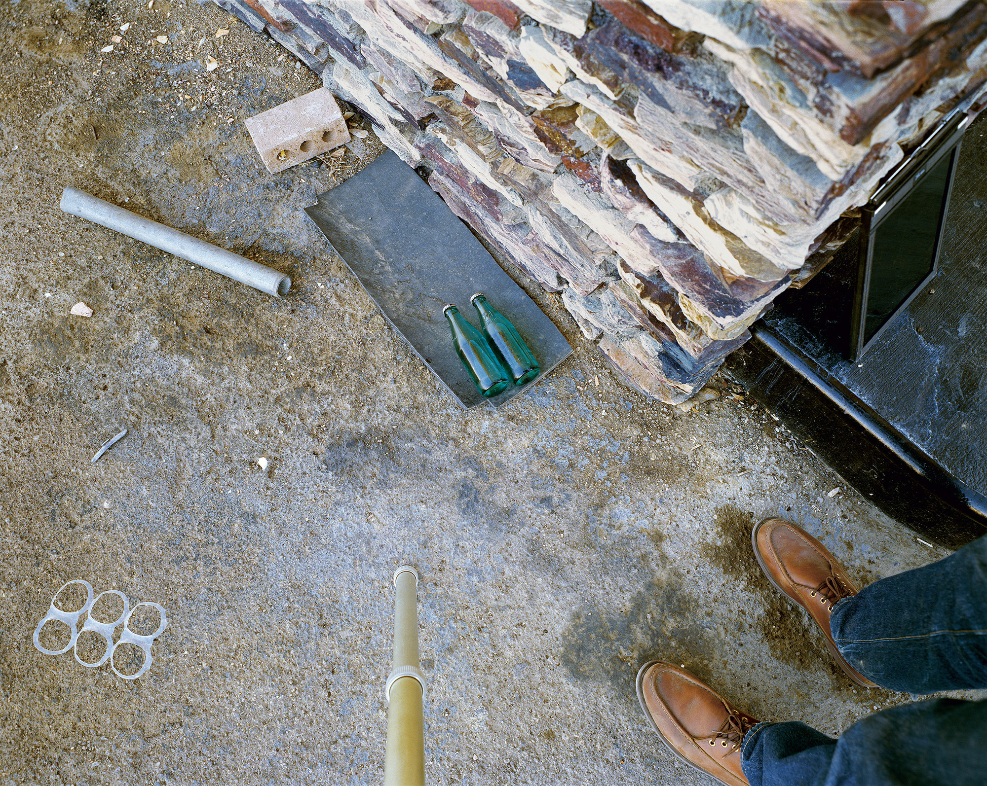 Various objects on the ground, including a plastic six-pack ring, bottles, and a stack of stonework, are shown next to a person&#x27;s feet 