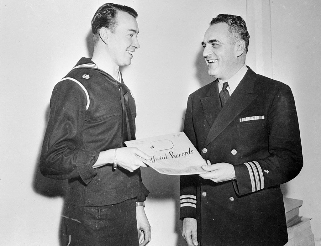 Young man in uniform smiles at an older man in uniform as they both hold an &quot;Official Records&quot; sheet of paper