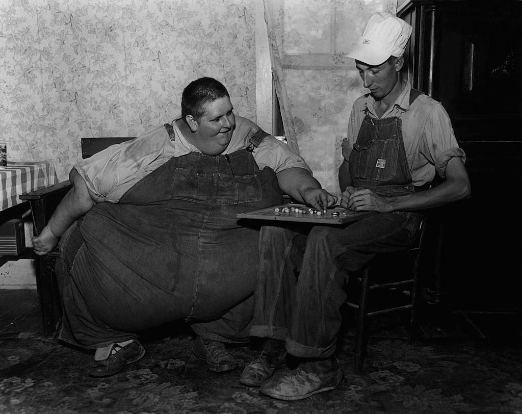 Black-and-white photo of very large man in overalls sitting and playing a board game with another man