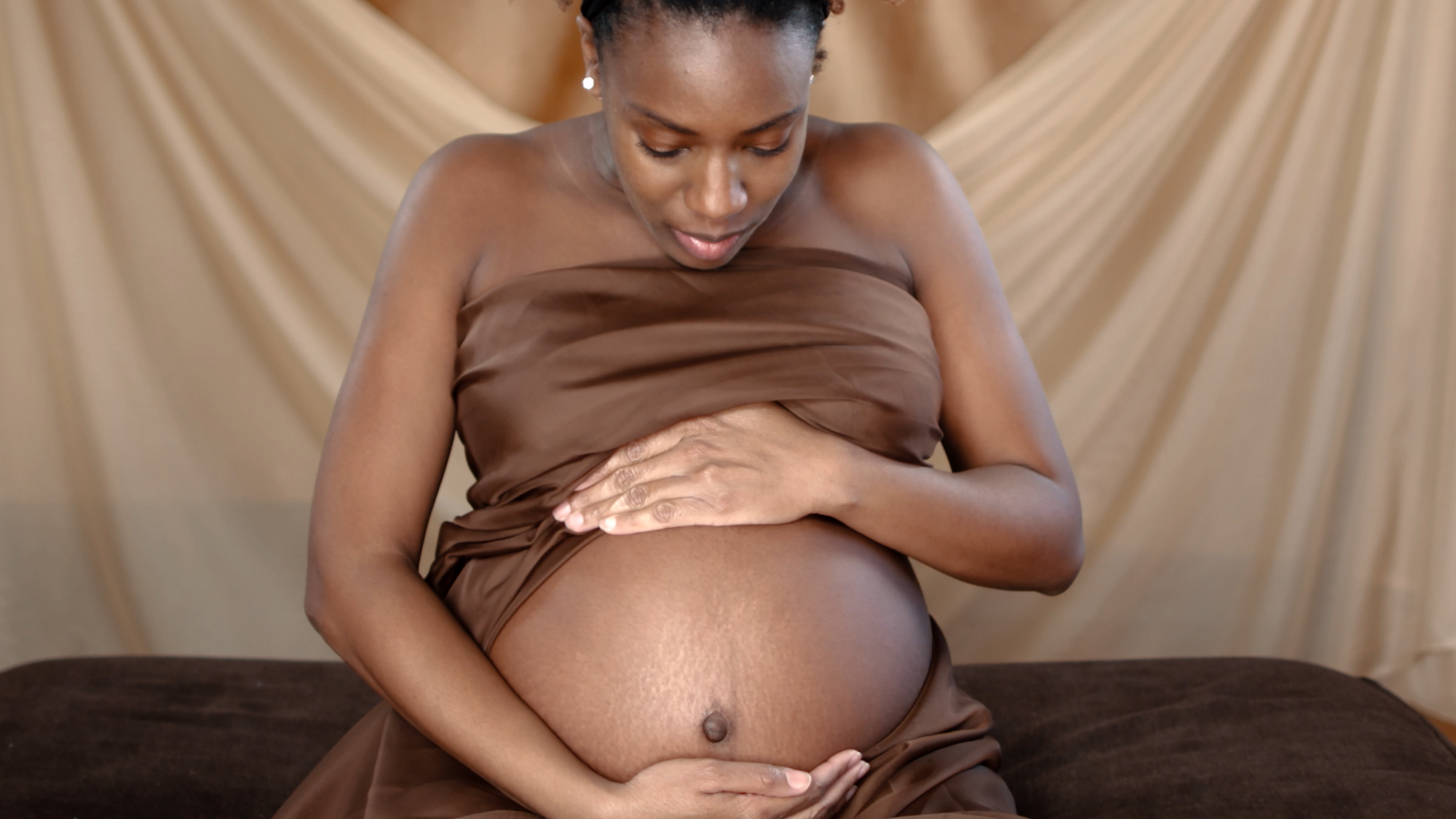 A Black, pregnant woman wrapped in brown sheets holds her bare stomach