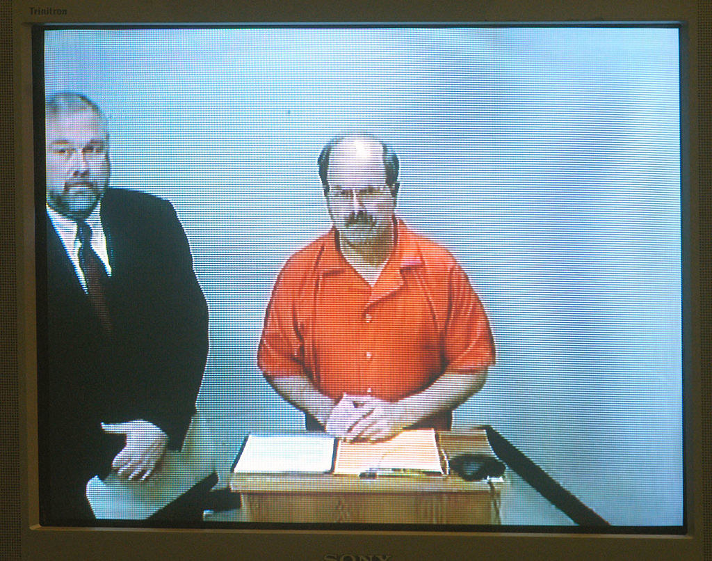 Rader in a prison jumpsuit standing at a podium next to a man in a suit and tie