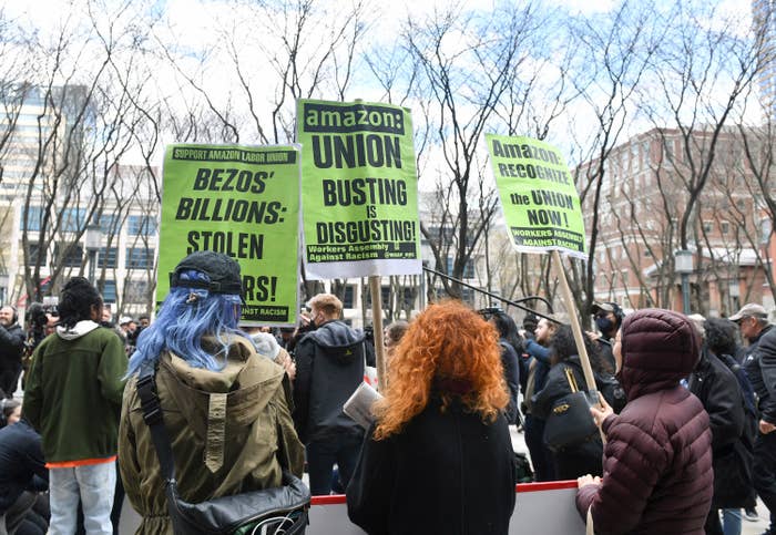 A crowd of protesters gather, some carrying signs like &quot;Amazon: Union Busting Is DisgustingQ&quot; and &quot;Recognize the Union Now!&quot; 