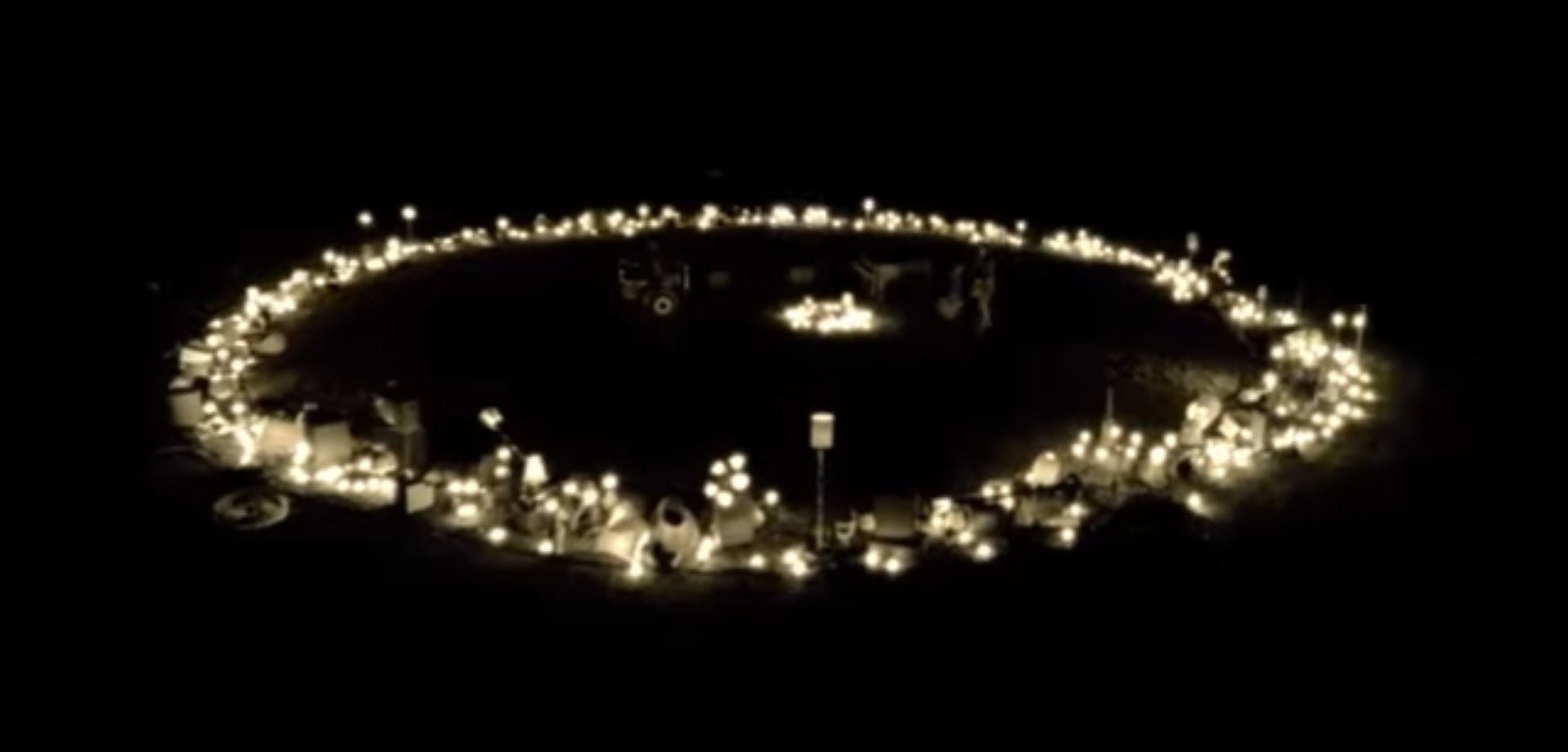 A circle formation of lights in a field at night. The band is barely visible, playing in the centre of the circle
