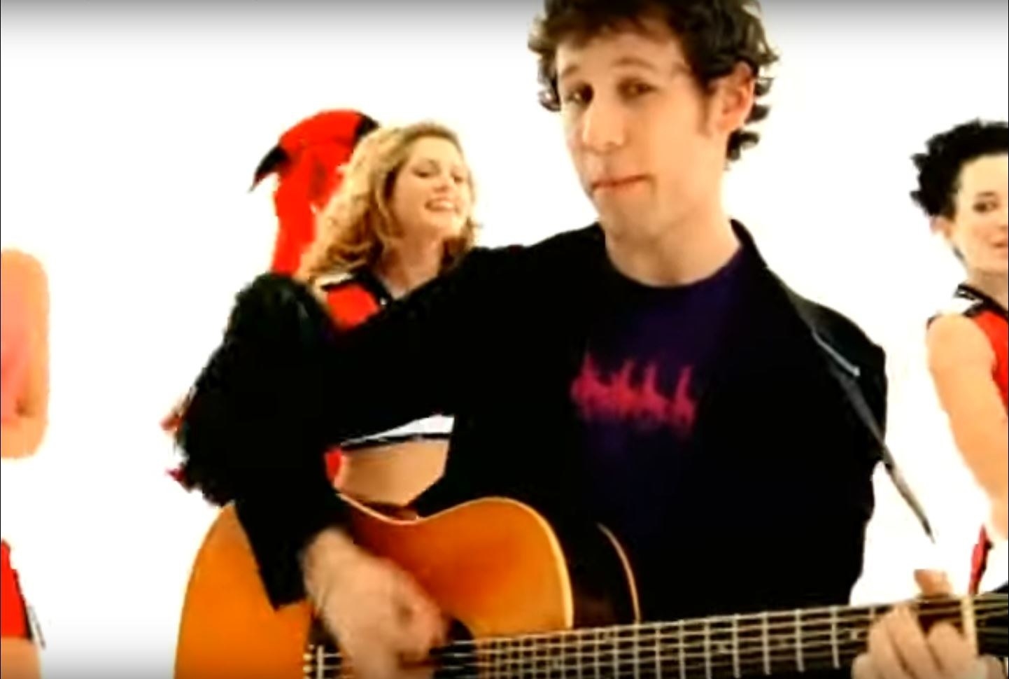 Ben Lee singing and playing his acoustic guitar in a white void as cheerleaders dance behind him