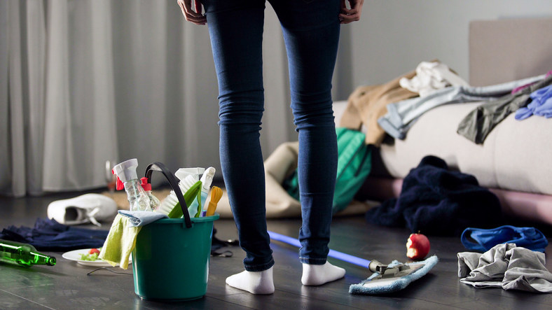 A person stands in a messy room with a bucket of cleaning products on the floor beside them