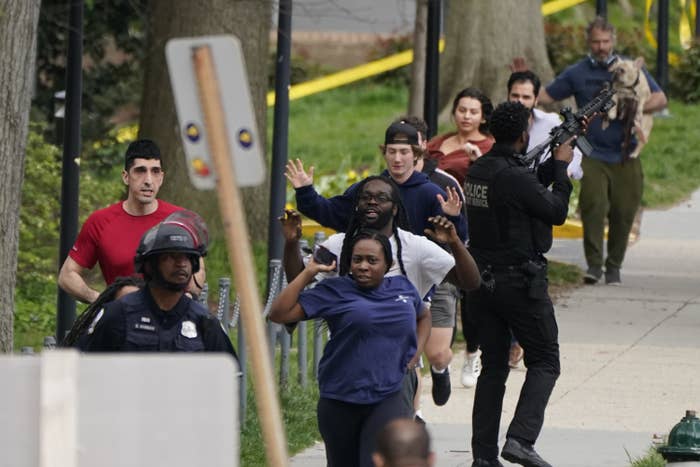 People, some with their arms up, run along the sidewalk as police officers stand nearby