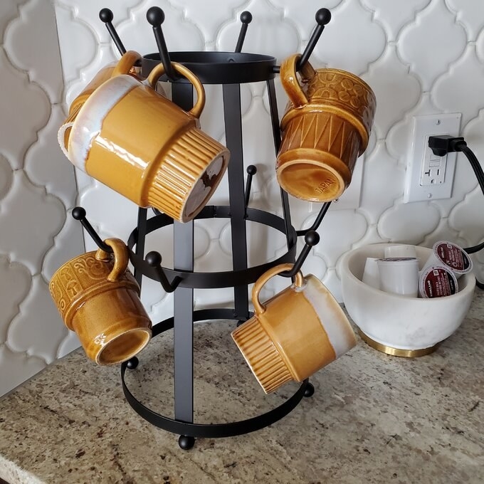 Four brown mugs are displayed on a metal mug tree on a kitchen counter, next to a bowl holding coffee pods