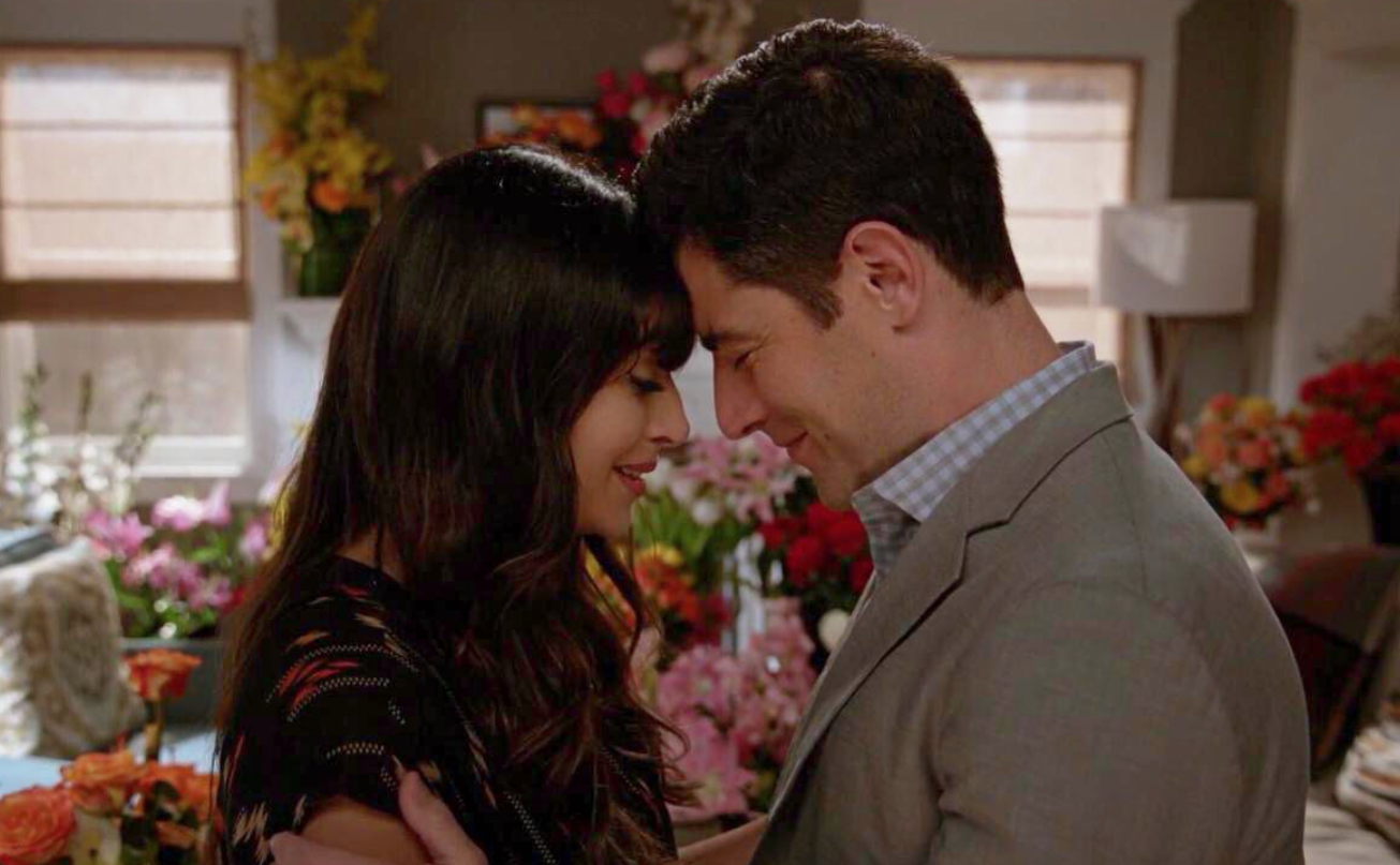 Cece and Schmidt lean their foreheads together while bouquets of flowers stand behind them