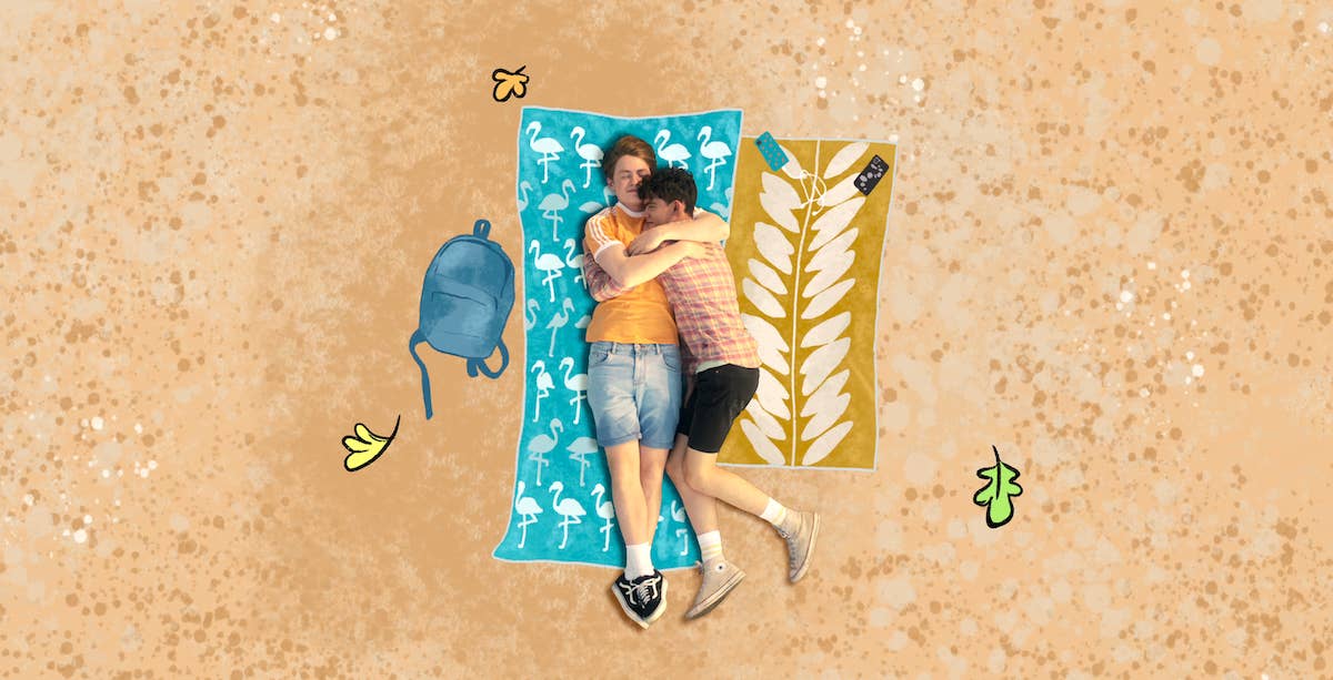 A still from Heartstopper shows two boys Nick and Charlie lying on the sand hugging each other with animated leaves from the comic around them
