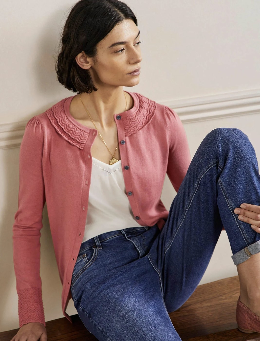 Model sitting against a wall wearing the pink button-up cardigan over a white tank