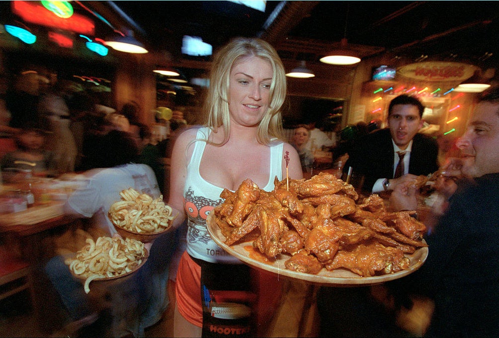 A Hooters waitress holding a tray of wings and plates of fries.