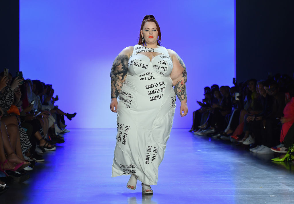 Tess Holliday walking the catwalk wearing a full white dress with &quot;Sample size&quot; printed all over it
