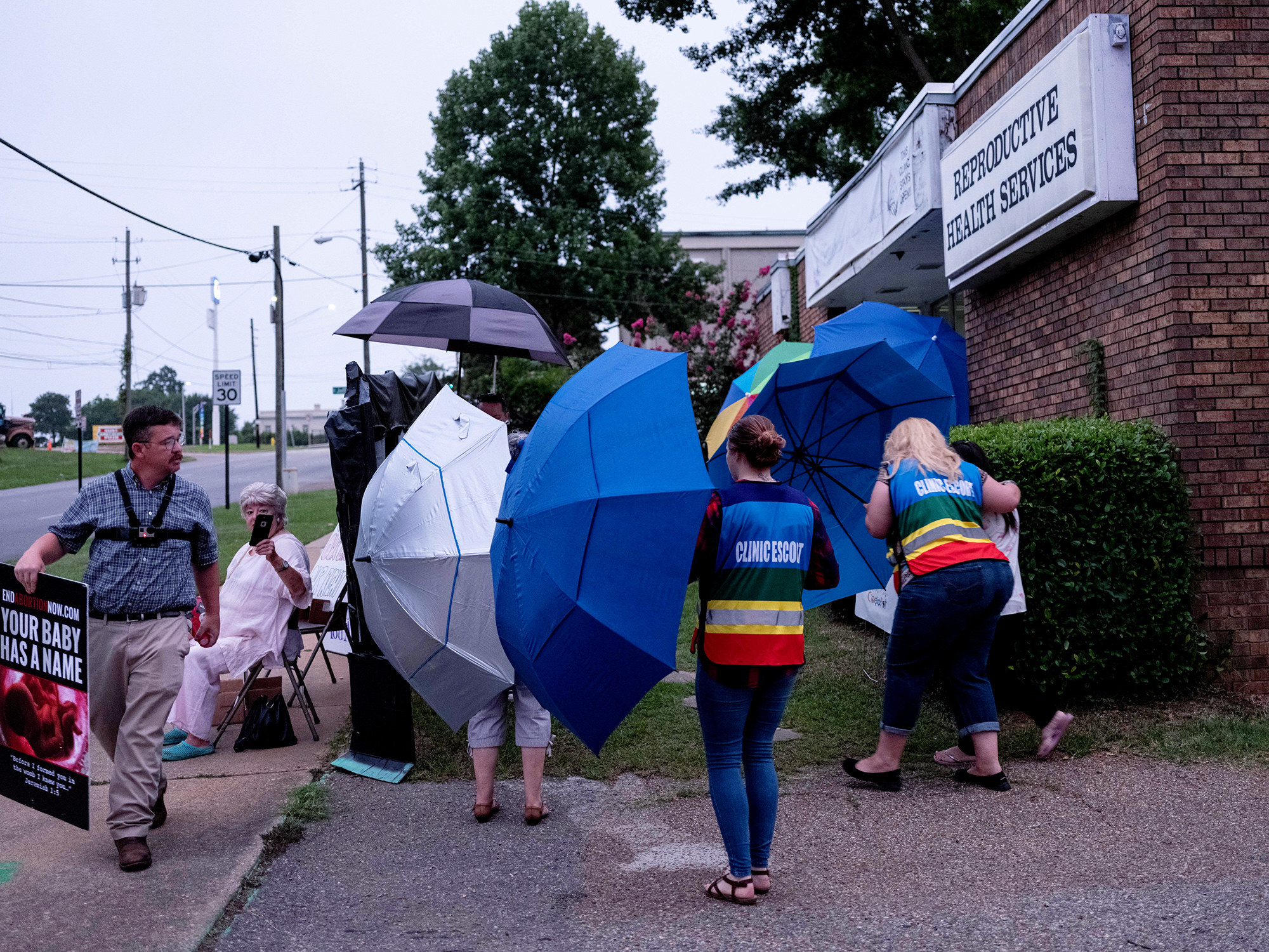 A group of people carry large umbrellas to shield patients from a man carrying a sign with a photo of a nearly full term fetus that says your baby has a name
