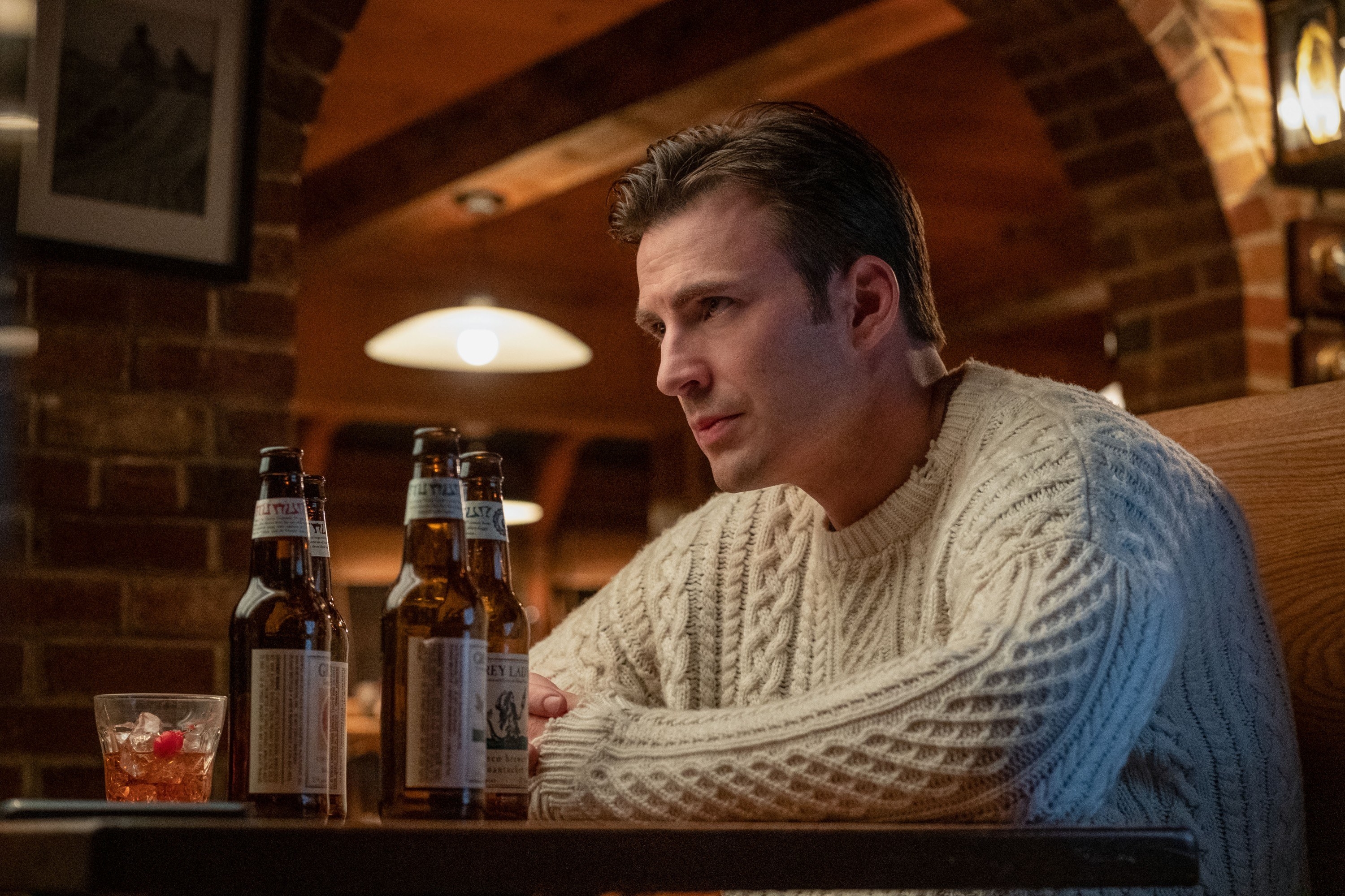 Chris Evans wearing a cream-colored sweater and sitting in a booth with beer bottles on the table