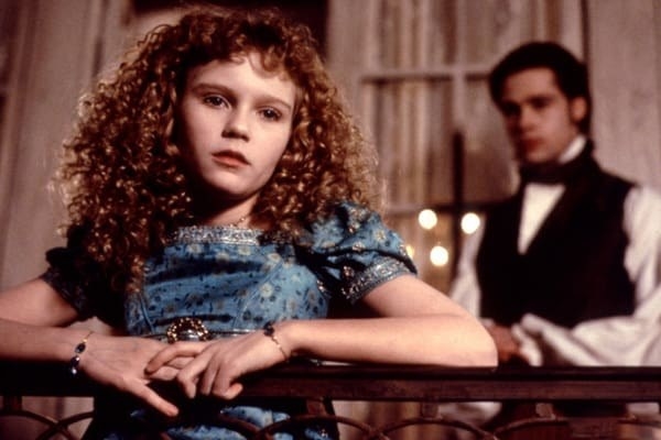 Kirsten, with long curls, leaning on a balcony with Brad Pitt in the background