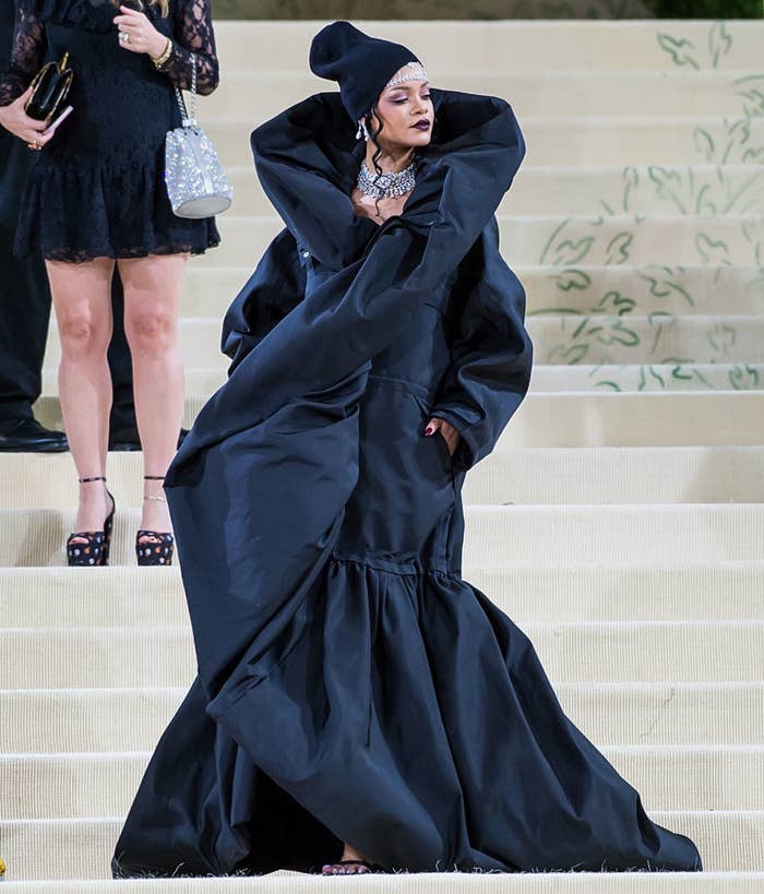 Rihanna wears a beanie and a long dramatic dress with pockets and tall collar