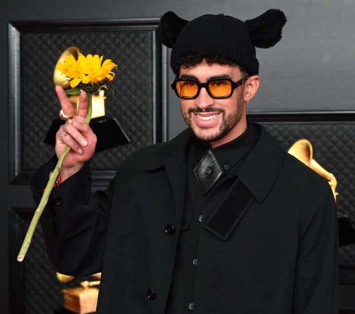 Bad Bunny smiling and giving the peace sign as he holds a sunflower at a red carpet event