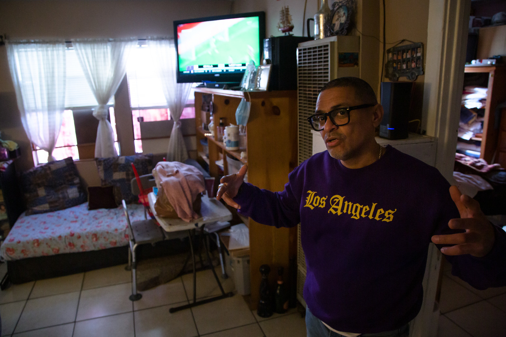 A man wearing thick-framed glasses gestures as he stands in a living room