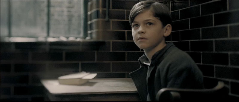 A young Tom Riddle in a dark room with black tiles, in front of a desk