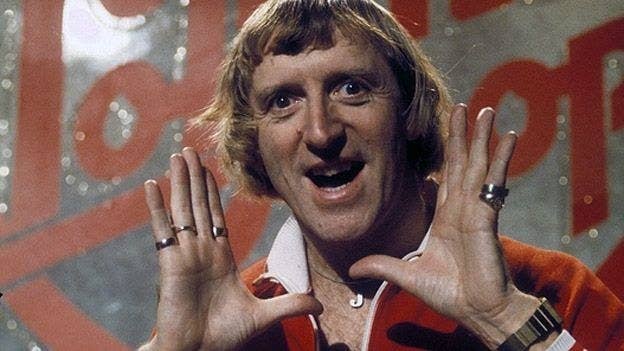 A picture of Jimmy Savile with his hand up near his face smiling happily into the camera